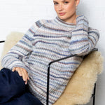 Woman sitting on a chair with a textured blanket wrapped around her, wearing a striped sweater and dark pants.
