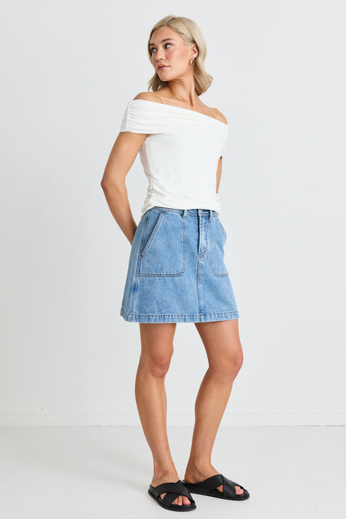 model wears an off the shoulder white top and a blue denim mini skirt and black sandals