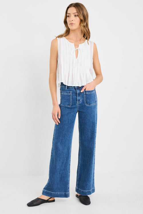 Woman wearing an ivory pleated sleeveless top with tie neckline, styled with high-waisted blue jeans.