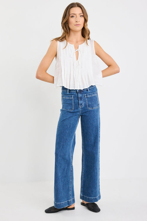 Woman wearing an ivory pleated sleeveless top with tie neckline, styled with high-waisted blue jeans.