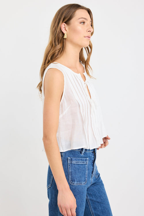 Woman wearing an ivory pleated sleeveless top with tie neckline, styled with high-waisted blue jeans.