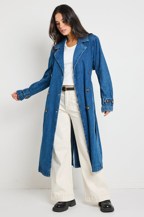 Model wears blue denim trench coat with tie waist and buttons