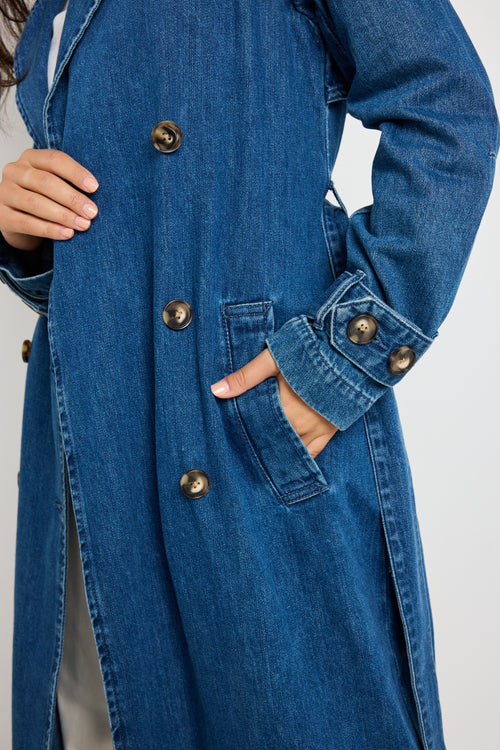 Model wears blue denim trench coat with tie waist and buttons