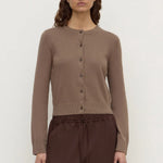 A woman modeling a taupe brown button-up cardigan sweater paired with dark brown drawstring trousers, shot against a plain light gray studio background.