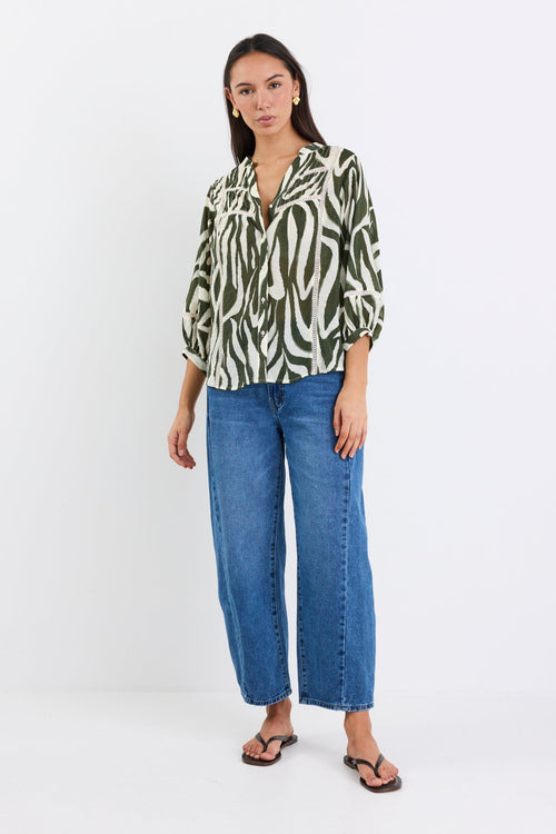 Woman wears a khaki and cream zebra-print shirt with lace inserts and pintuck detail, styled with gold earrings and blue jeans.