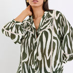 Woman wears a khaki and cream zebra-print shirt with lace inserts and pintuck detail, styled with gold earrings and blue jeans.