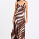 Woman wearing a chocolate brown satin midi dress with white polka dots, adjustable spaghetti straps, and a bias-cut silhouette, standing against a white background.