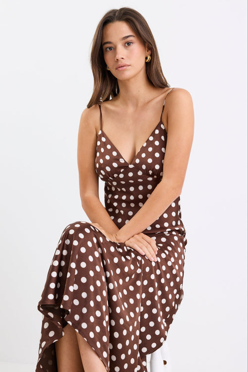 Woman wearing a chocolate brown satin midi dress with white polka dots, adjustable spaghetti straps, and a bias-cut silhouette, standing against a white background.