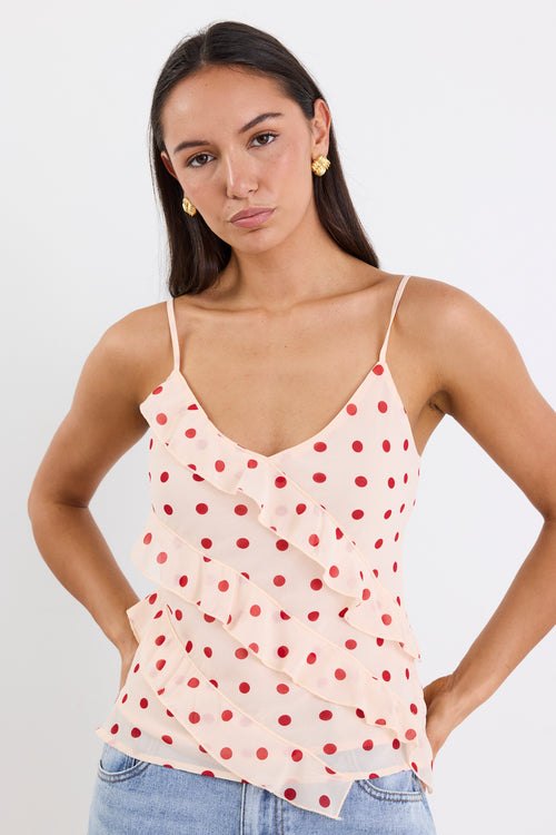 Woman wearing a cream cami top with cherry-red polka dots, ruffled front detailing, and thin adjustable straps, paired with light blue jeans against a white background.