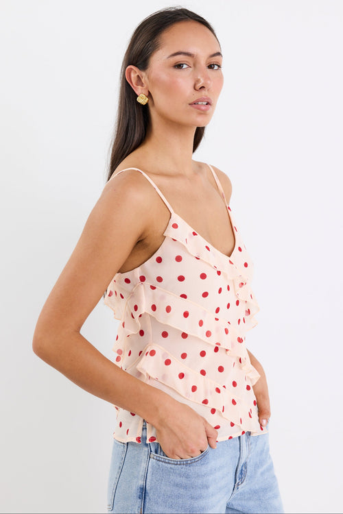 Woman wearing a cream cami top with cherry-red polka dots, ruffled front detailing, and thin adjustable straps, paired with light blue jeans against a white background.