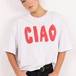 A woman wearing a white oversized t-shirt with the word 'CIAO' printed in red across the chest, paired with black jeans and a gold-buckled belt, posing against a plain white wall.