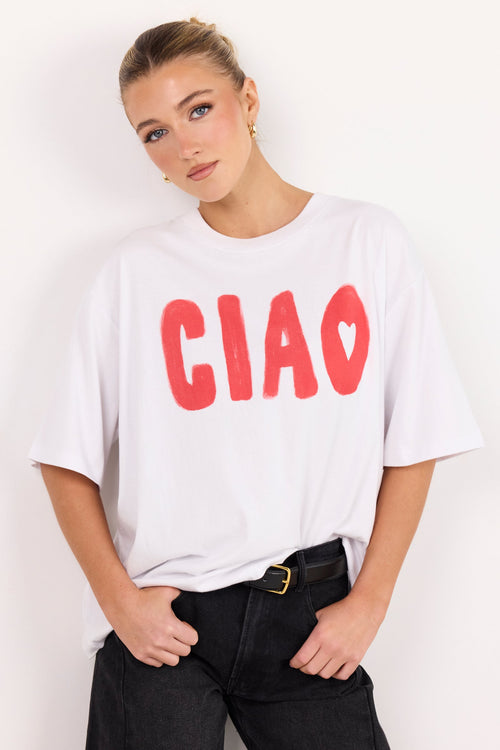 A woman wearing a white oversized t-shirt with the word 'CIAO' printed in red across the chest, paired with black jeans and a gold-buckled belt, posing against a plain white wall.