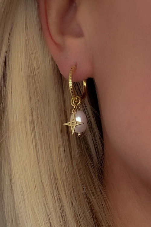 Close-up of a gold sleeper earring worn on a woman's ear, featuring a hanging pearl and gold star charm against blonde hair.
