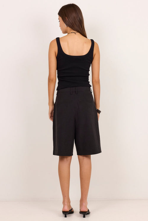 A woman is shown from the back wearing a black sleeveless tank top and black wide-leg shorts, standing against a plain off-white studio background.