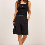 A woman modeling a black sleeveless tank top and black wide-leg shorts with a matching black belt, standing against a plain off-white studio background.