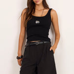 A woman modeling a black sleeveless tank top, black wide-leg shorts, a black belt, and black sandals against a plain off-white studio background.