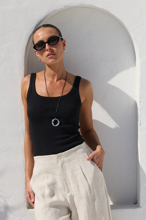 A woman wearing a black ribbed tank top, wide-leg cream linen trousers, and black sunglasses stands in front of a white stucco wall with an arched niche.