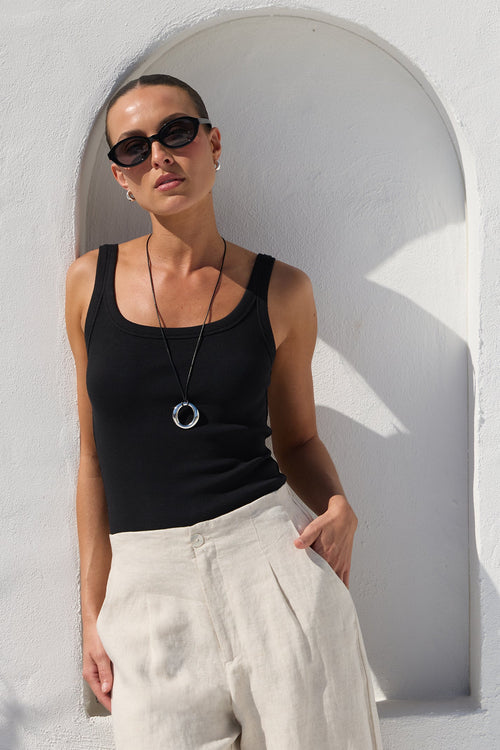 A woman wearing a black ribbed tank top, cream wide-leg linen trousers, black sunglasses, and a silver ring pendant necklace, posing against a white stucco wall with an arched niche.