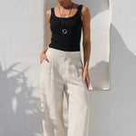 A woman wearing a black sleeveless tank top and cream wide-leg linen trousers stands in a sunlit white-walled courtyard next to a large terracotta planter with a palm plant.