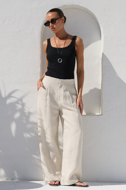 A woman wearing a black sleeveless tank top and cream wide-leg linen trousers stands in a sunlit white-walled courtyard next to a large terracotta planter with a palm plant.
