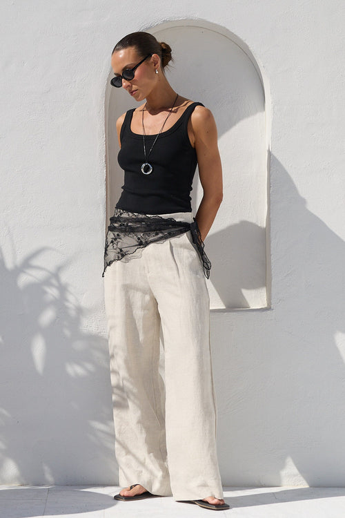 A woman wearing a black tank top, wide-leg cream trousers, and a black lace sash stands against a white stucco wall with a large potted palm plant to her left.