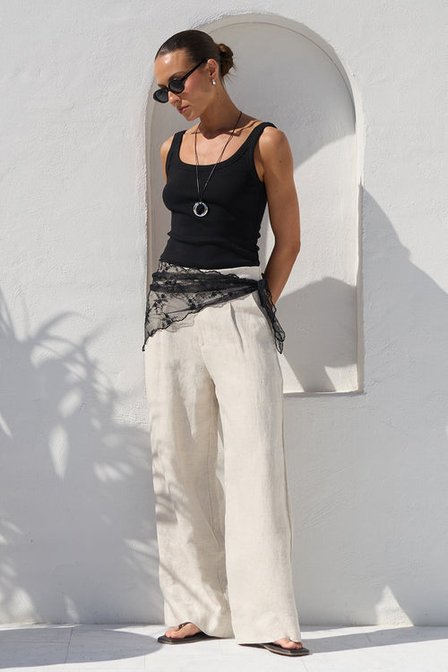 A woman wearing a black tank top, wide-leg cream trousers, and a black lace sash stands against a white stucco wall with a large potted palm on the left.