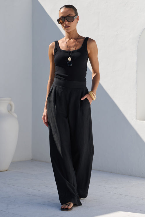 A woman modeling a black sleeveless top and wide-leg black pants in an outdoor setting with white stucco walls and strong sunlight.