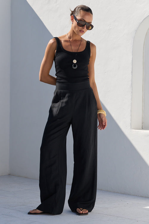 A woman wearing a black sleeveless top and matching wide-leg pants stands in a minimalist white architectural setting with strong sunlight and shadows.
