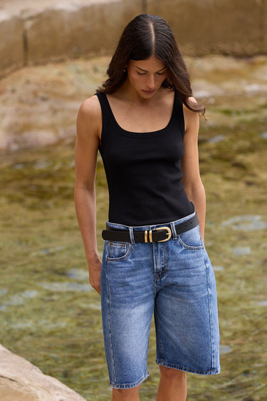 A woman standing outdoors on a stone ledge next to a shallow stream, wearing a black sleeveless top, blue denim cutoff shorts, a black belt with a gold buckle, and black flip-flops.