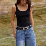 A woman standing on a stone ledge next to a shallow stream, wearing a black tank top, blue denim cutoff shorts, a black belt with a gold buckle, and black flip-flops.