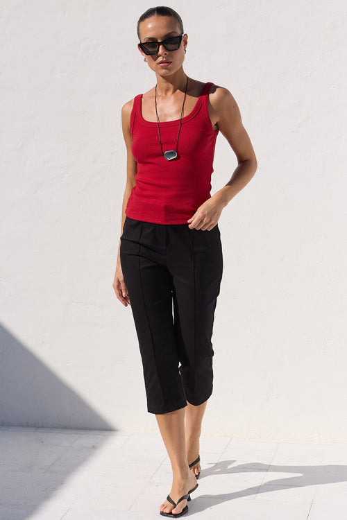 A woman modeling a bright red sleeveless tank top and black cropped capri pants against a minimalist white wall background.