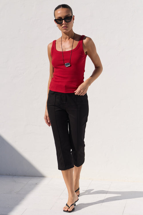 A woman modeling a bright red sleeveless tank top and black cropped capri pants against a minimalist white wall background.