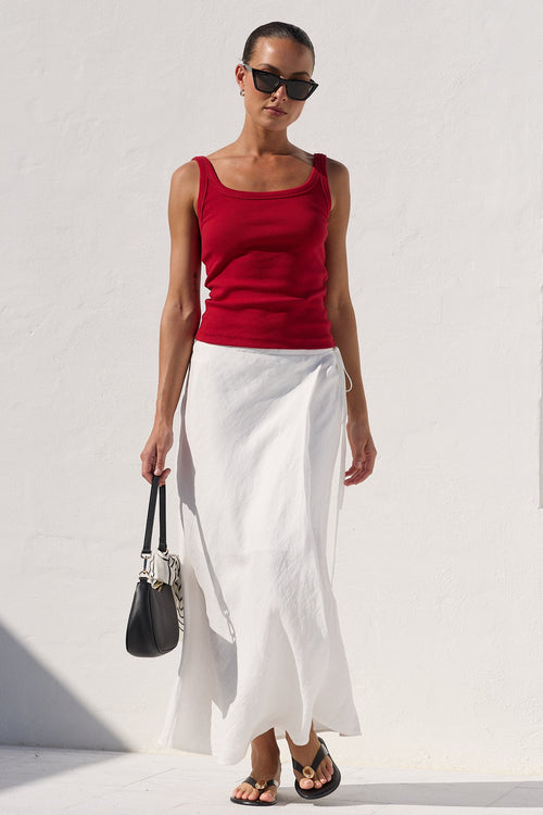 A woman stands outdoors against a bright white wall wearing a sleeveless red top and a long white skirt, holding a black handbag.