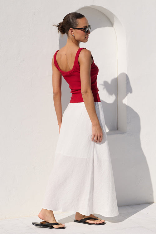 A woman stands in profile against a bright white wall with a recessed arch detail, wearing a fitted red tank top, a long white skirt, and black flip-flop sandals.