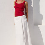 A woman wearing a red ribbed tank top and white wide-leg skirt or maxi skirt, styled with black sunglasses and black flip-flops, posing against a white stucco wall with a strong arch shadow.