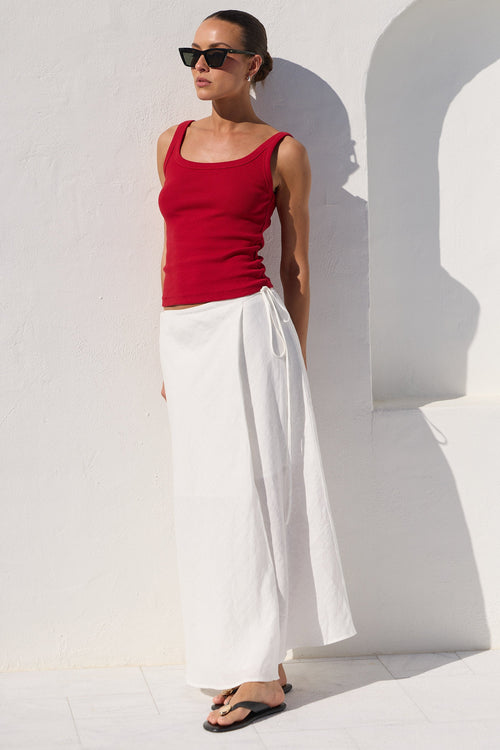 A woman wearing a red ribbed tank top and white wide-leg skirt or maxi skirt, styled with black sunglasses and black flip-flops, posing against a white stucco wall with a strong arch shadow.