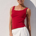 A woman wearing a red tank top and white wide-leg linen pants stands against a white stucco wall with a recessed arch, casting a shadow.
