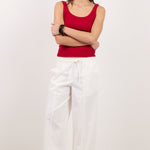 A woman stands against a plain white studio background wearing a sleeveless red tank top, loose white drawstring pants, and black flip-flop sandals, with her arms crossed.
