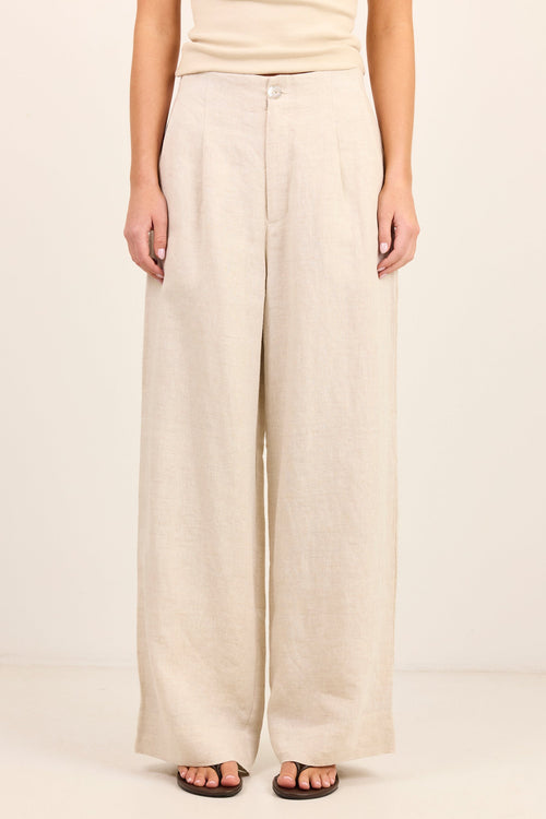 A model wearing a pair of wide-leg linen pants in a light beige color, paired with a matching beige top and dark brown sandals, set against a plain cream background.