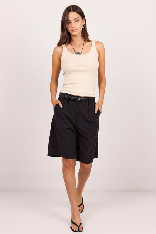 A woman modeling a pair of black wide-leg shorts with a black belt, paired with a cream-colored sleeveless top and black sandals, set against a plain light gray studio background.