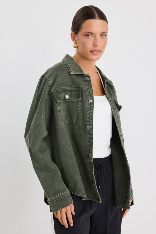 Woman wearing a khaki green washed denim jacket with silver buttons, styled with black pants featuring a cream side stripe.





