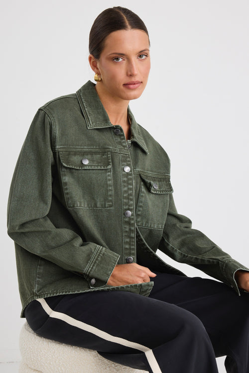 Woman wearing a khaki green washed denim jacket with silver buttons, styled with black pants featuring a cream side stripe.





