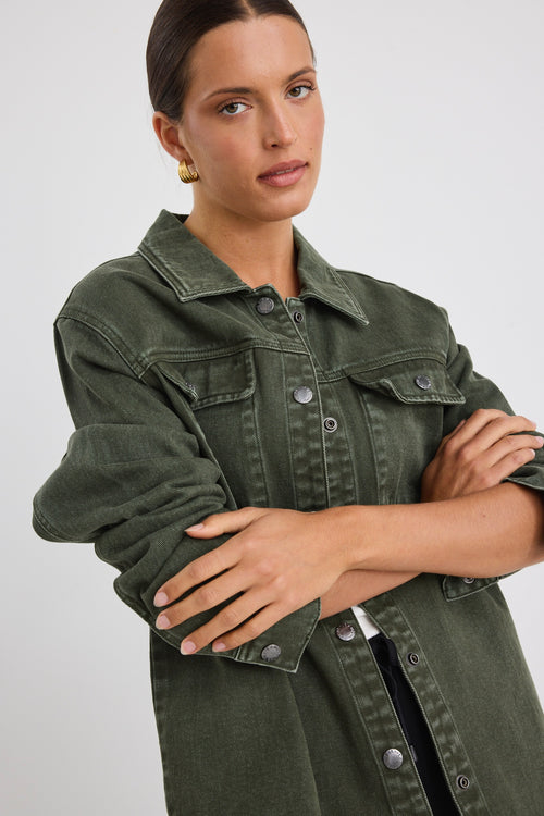 Woman wearing a khaki green washed denim jacket with silver buttons, styled with black pants featuring a cream side stripe.
