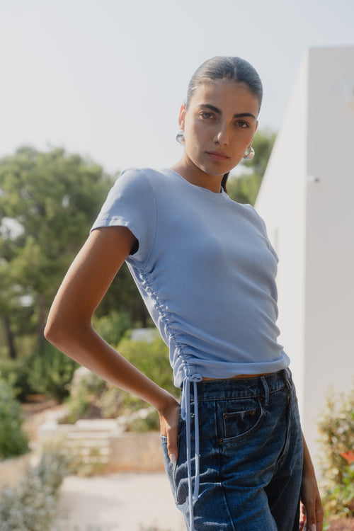 Person wearing a light blue t-shirt and jeans outdoors with greenery in the background