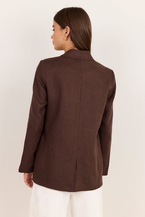 A woman shown from the back wearing a dark brown tailored blazer and white pants, photographed against a plain light beige studio background.