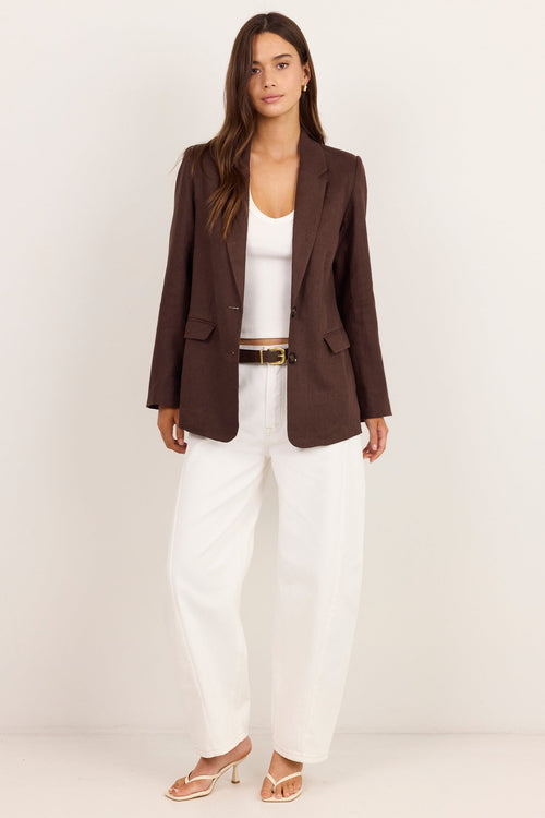 A woman modeling a dark brown blazer over a white tank top and white wide-leg trousers, paired with a thin brown belt and beige heeled sandals, set against a plain off-white studio background.