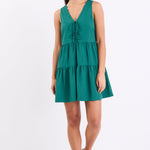 Woman wearing a forest green sleeveless mini dress with a v-neckline, front tie detail, and tiered skirt, standing against a white background.
