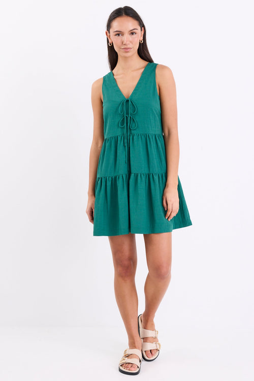 Woman wearing a forest green sleeveless mini dress with a v-neckline, front tie detail, and tiered skirt, standing against a white background.