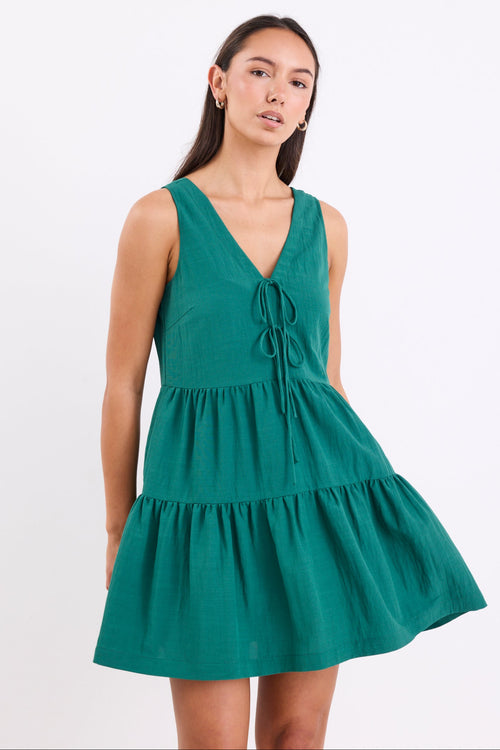Woman wearing a forest green sleeveless mini dress with a v-neckline, front tie detail, and tiered skirt, standing against a white background.