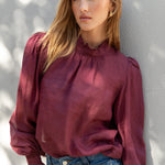 Woman wearing a burgundy blouse and blue jeans against a gray wall.